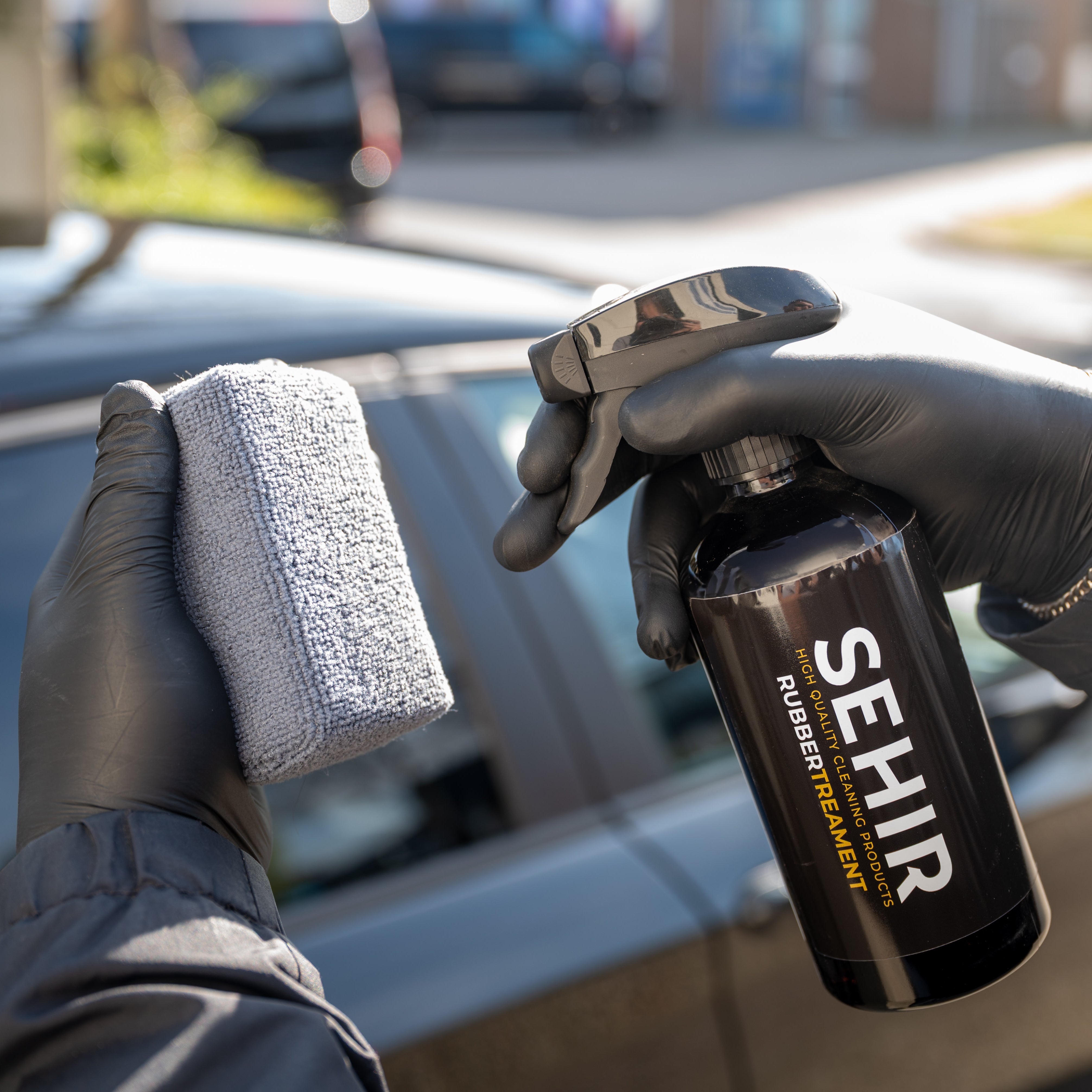 Person holding a spray bottle labeled 'SEHIR' and a sponge in front of a car.