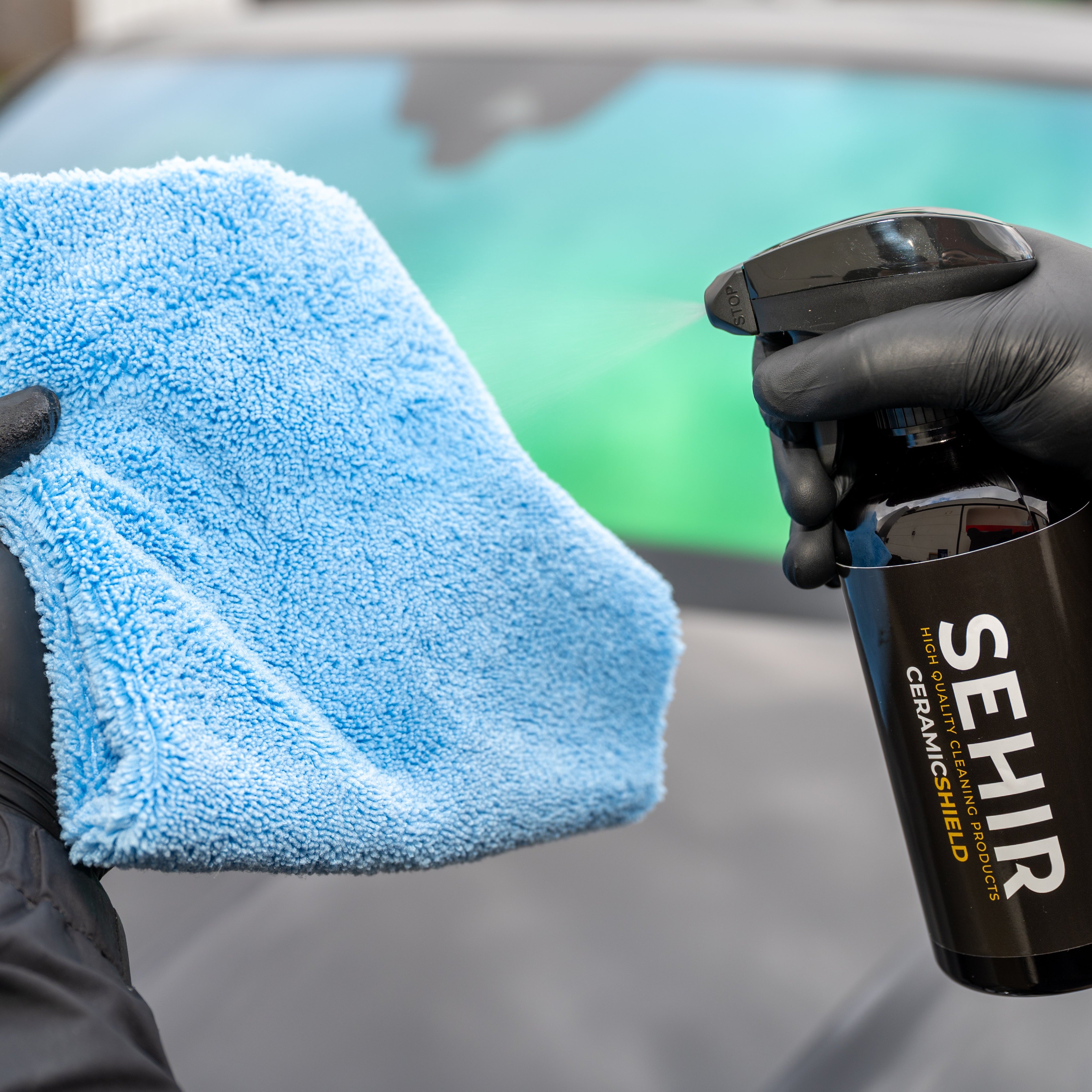 Person holding a blue microfiber cloth and a black spray bottle labeled 'SEHIR' in front of a car.