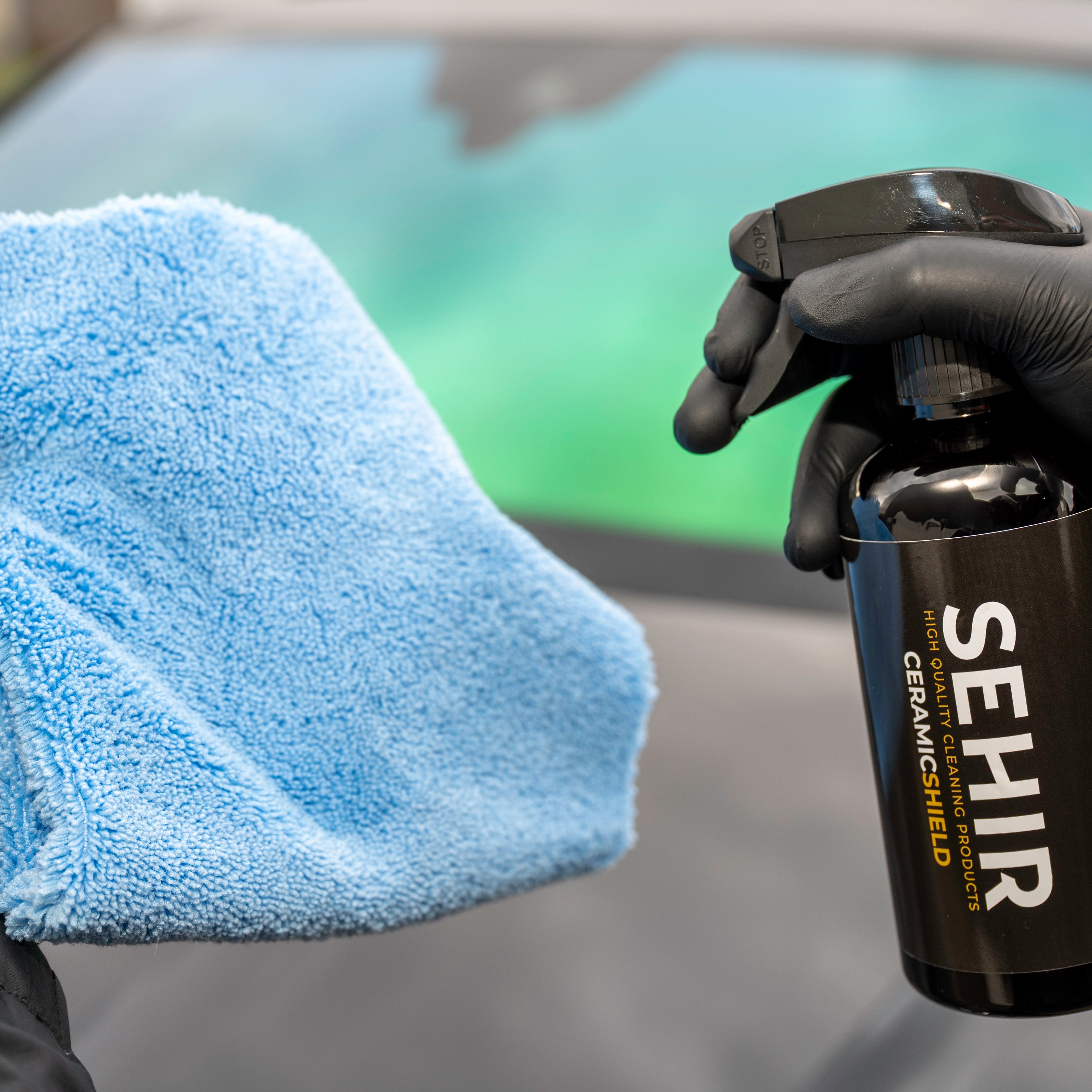 Person holding a blue microfiber cloth and a black spray bottle labeled 'SEHIR' in front of a car.