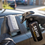 Person holding a spray bottle labeled 'SEHIR' and a sponge in front of a car.