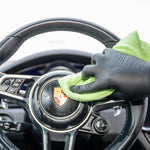 Hand wearing a black glove cleaning a car steering wheel with a green cloth.
