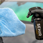 Person holding a blue microfiber cloth and a black spray bottle labeled 'SEHIR' in front of a car.